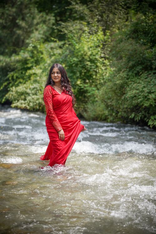 Free A woman in a red dress stands in a forest stream, surrounded by lush greenery. Stock Photo
