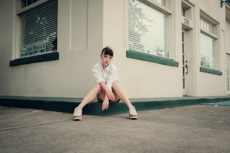 Woman In White Shirt Sitting In Building Corner