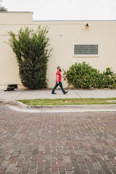 A man in a red shirt walks past a beige wall with lush greenery on a cobblestone street.