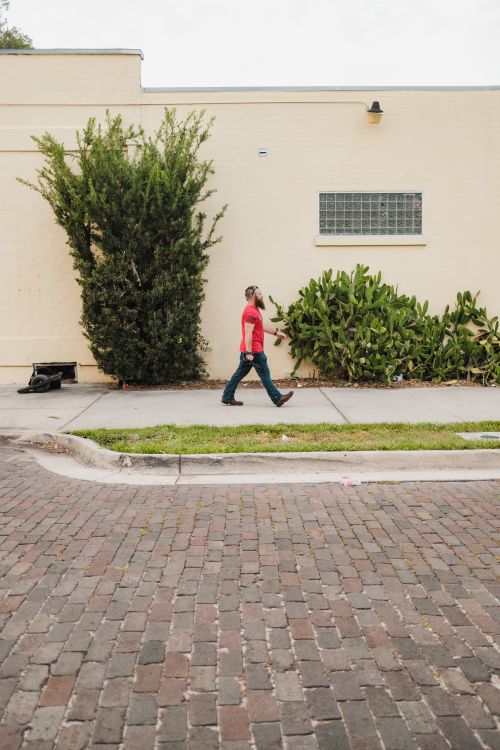 Free A man in a red shirt walks past a beige wall with lush greenery on a cobblestone street. Stock Photo