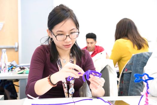 Young Asian woman engaged in craft activity in a classroom environment, creating with purple string.