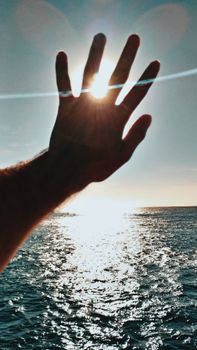 A hand silhouetted by the bright sun over a shimmering ocean during sunset, evoking tranquility and wonder.