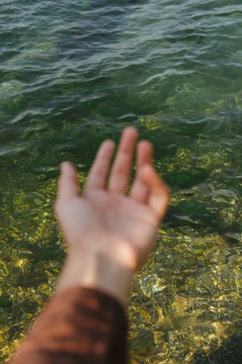 Free A hand reaching out towards clear, shimmering sea water on a sunny day. Stock Photo