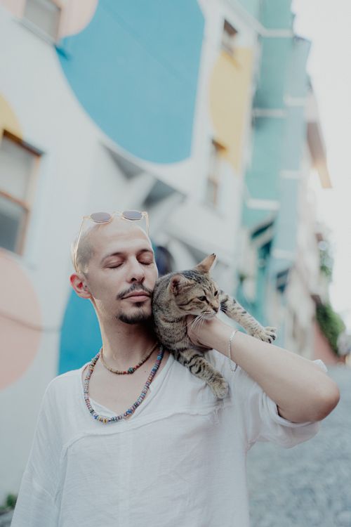 Free Man with sunglasses and a cat on shoulder, standing against colorful urban mural. Relaxed and stylish atmosphere. Stock Photo
