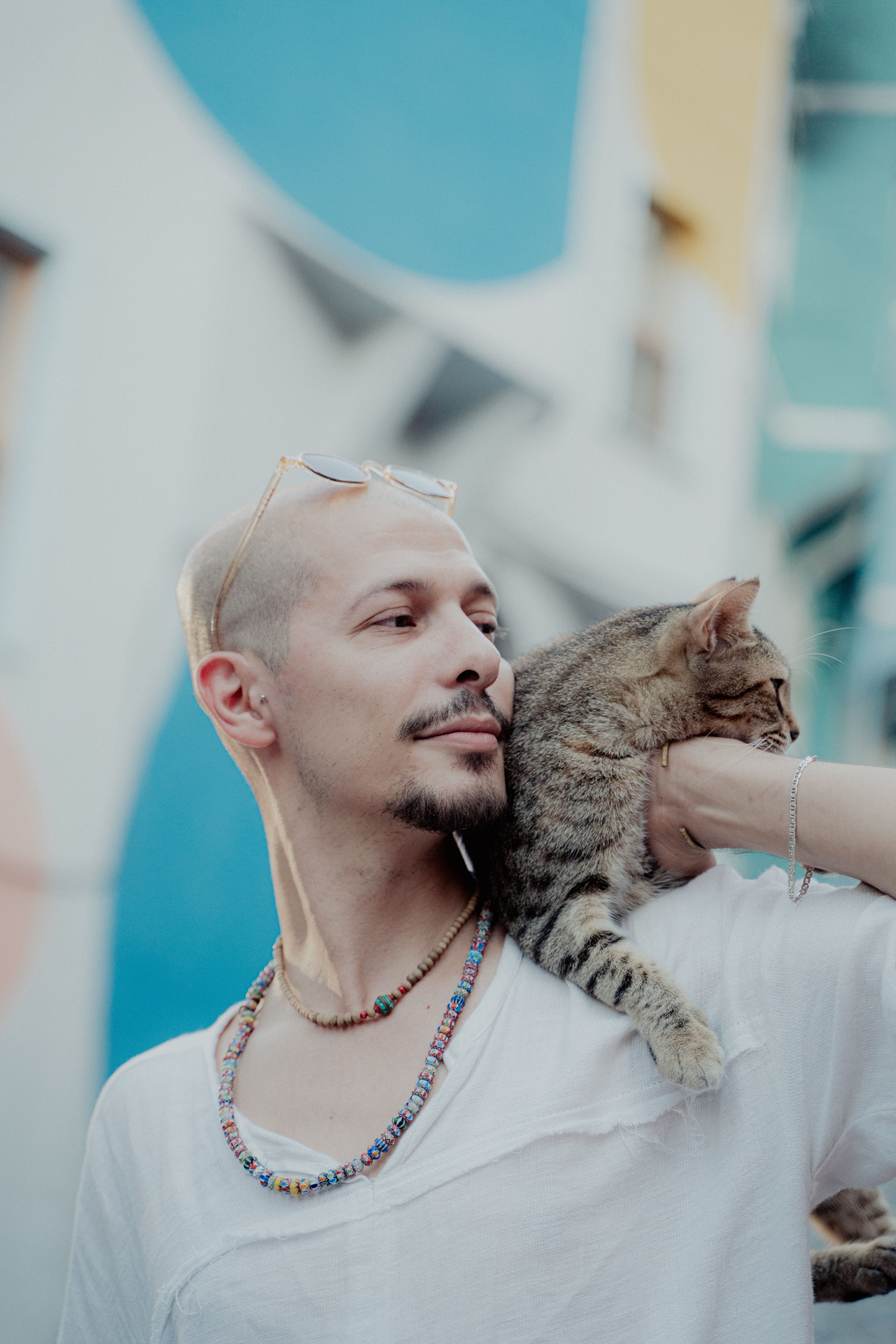 Man smiling with a tabby cat on his shoulder against a colorful urban backdrop.
