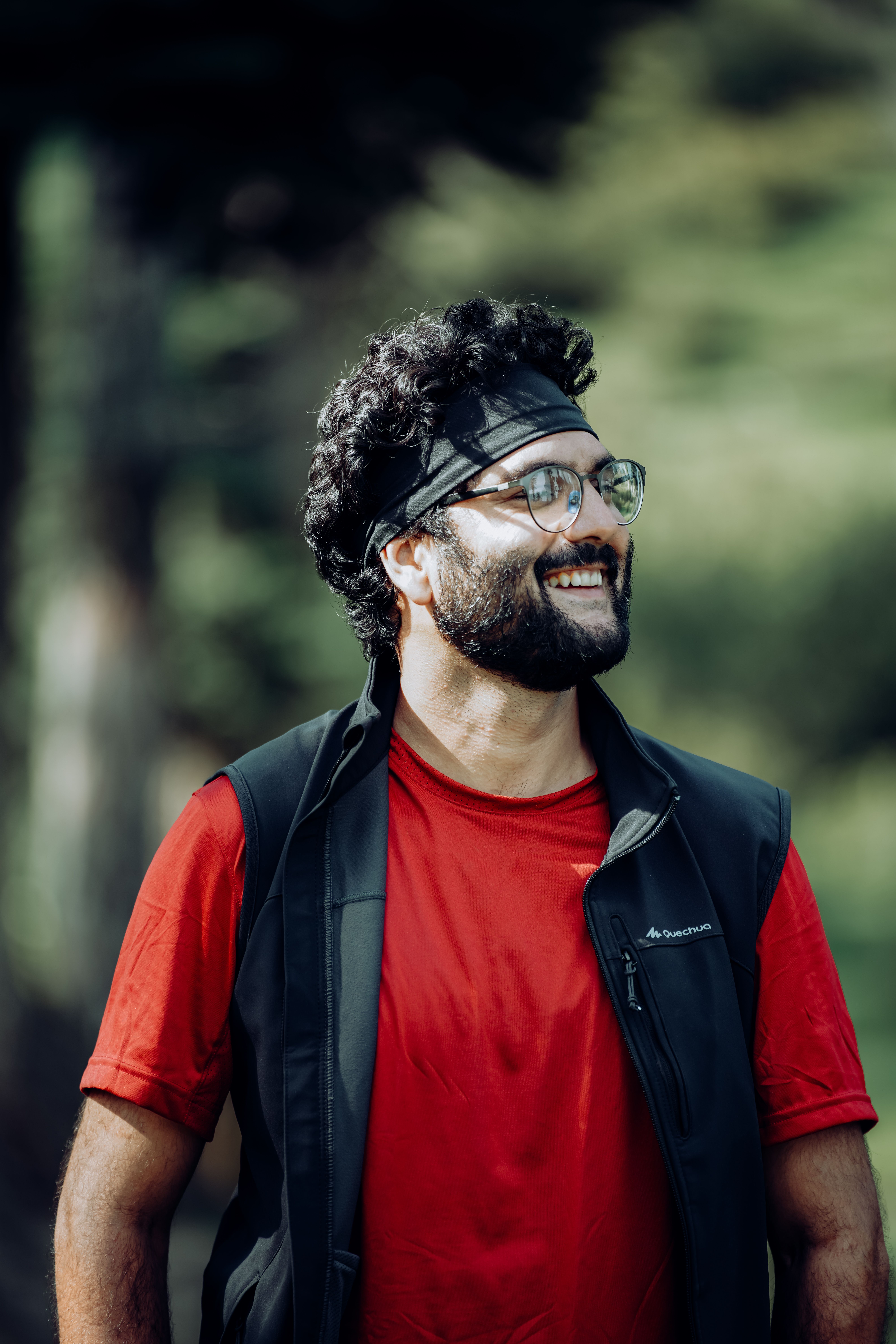Portrait of a smiling man in a red t-shirt and black vest outdoors with a blurred green background.