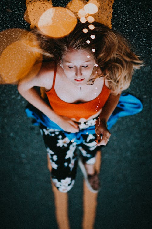 Photo of Woman in Tank Top Sleeping on Tarmac Road Holding String Lights