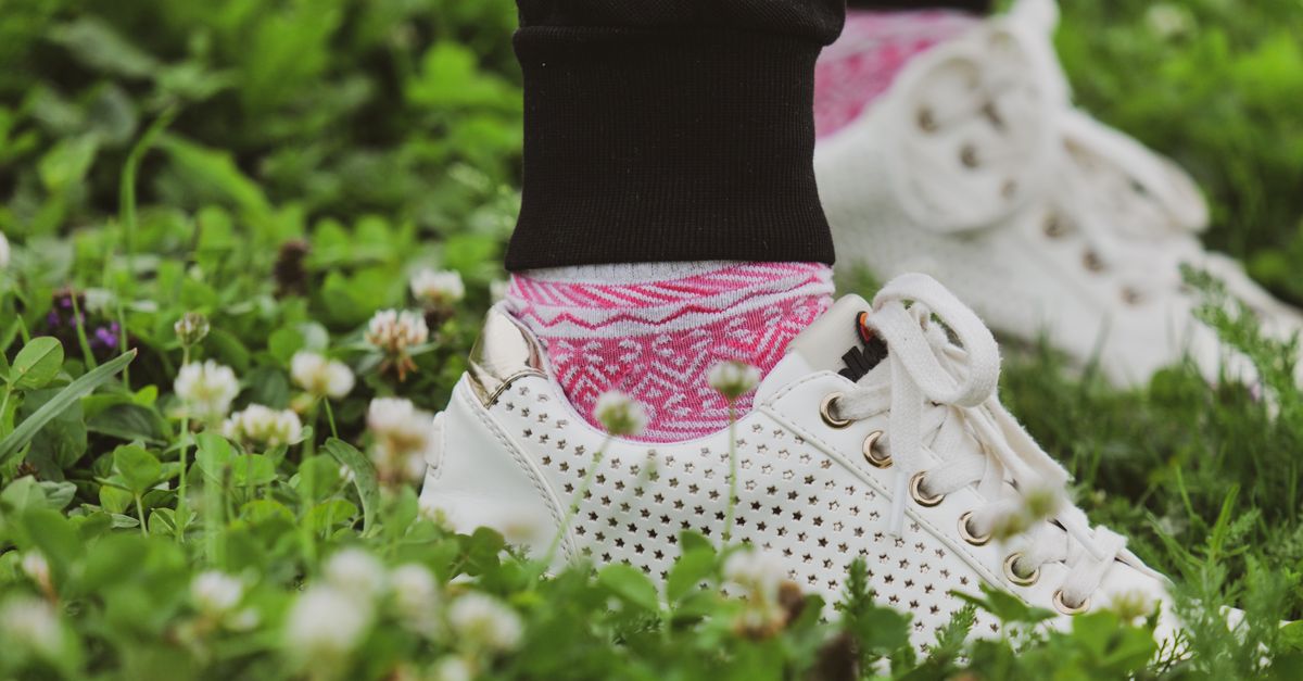 Close-up of white sneakers with pink socks amidst lush green grass and flowers.