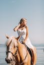 在一个阳光明媚的日子里，一位穿着白色裙子的年轻女子在海边骑着一匹金色的马