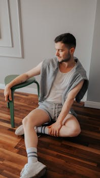 Young man in casual attire sits comfortably on a wooden floor indoors, exuding relaxation.