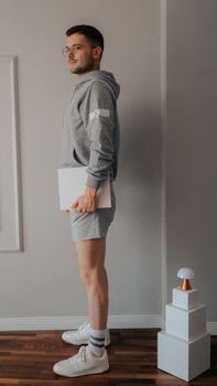 Man in gray hoodie holding a laptop, standing in a minimalistic room.