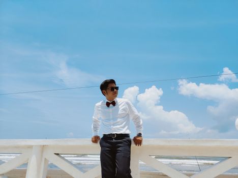 Well-dressed man in sunglasses lean against beach railings in Ilocos Region, Philippines under a blue sky.