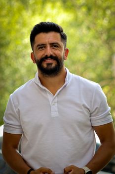 Smiling man with black hair and beard outdoors in a white polo shirt. Bright and cheerful portrait.