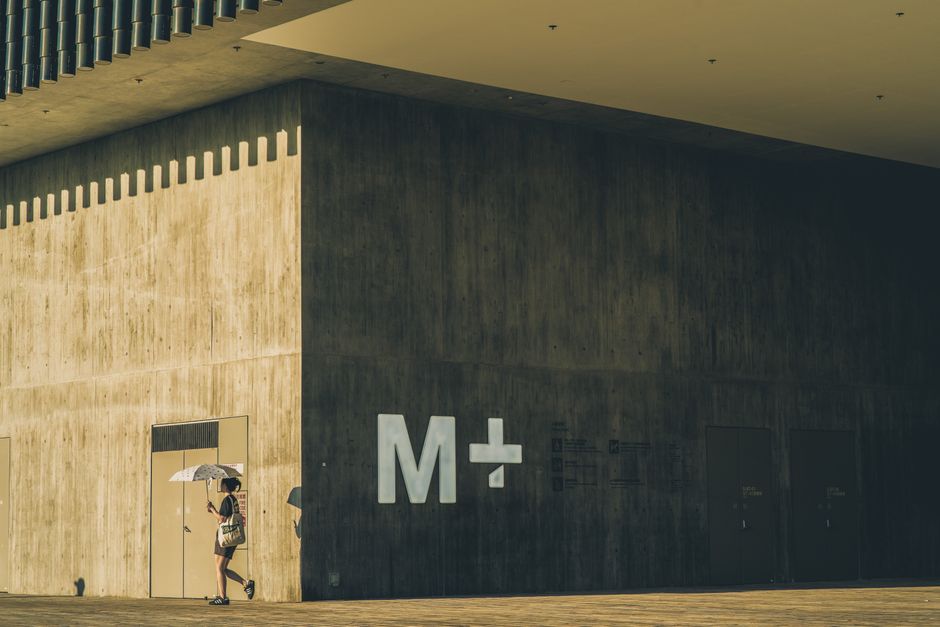 A woman walks by a modern concrete building with an umbrella on a sunny day.