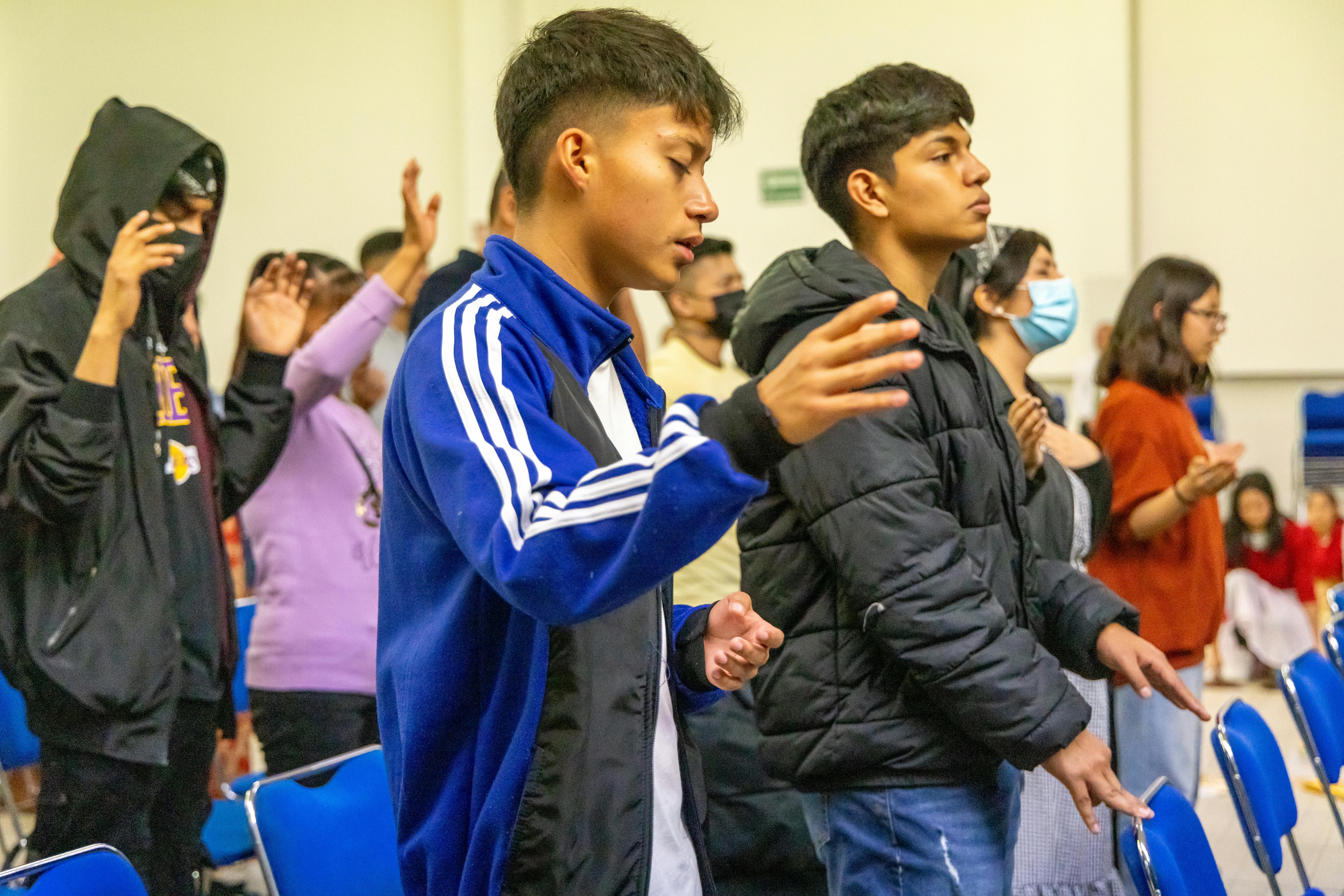 A group of teenagers attending a worship session indoors, showing diverse expressions of prayer.