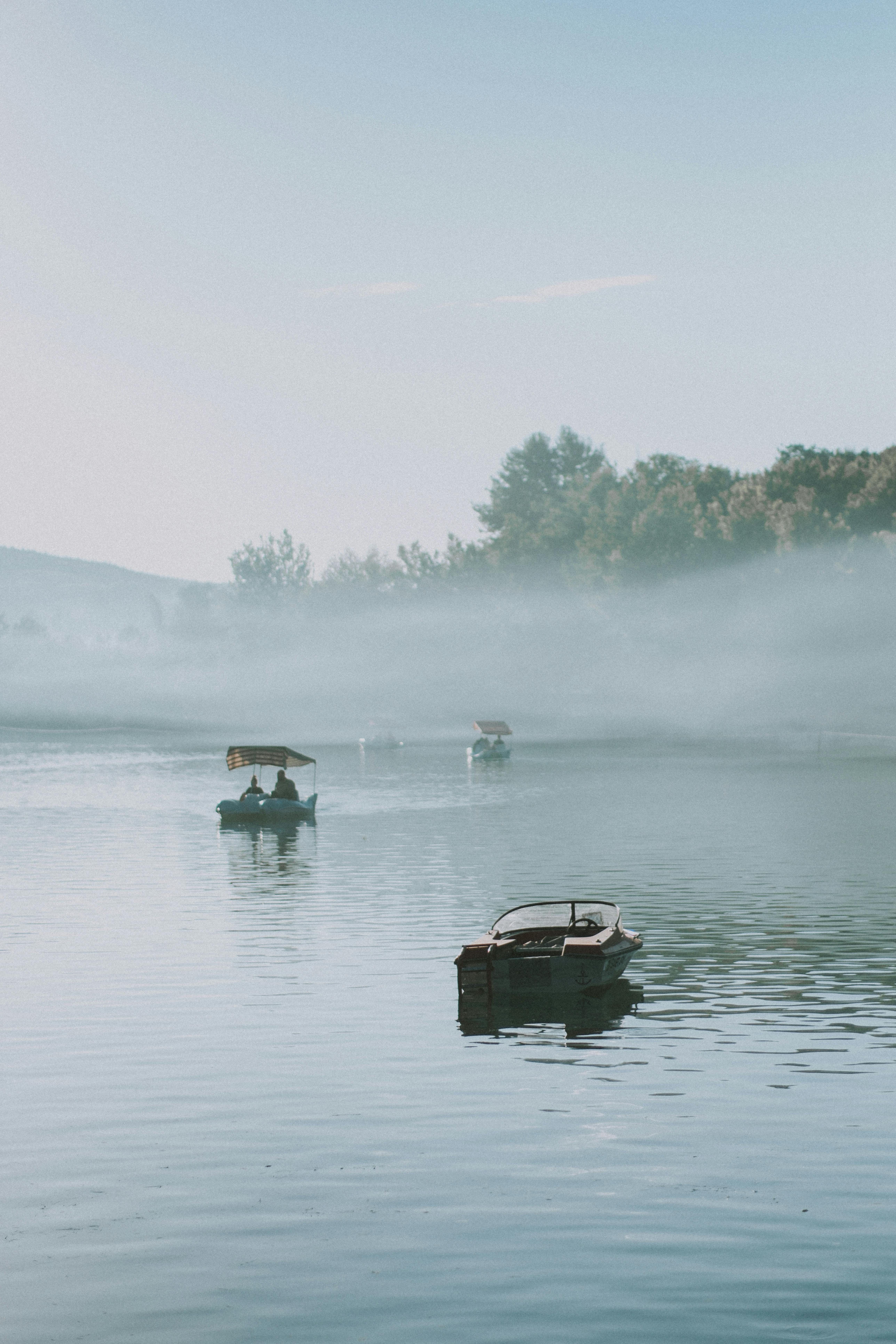 Tranquil scene of boats on a misty lake during morning, surrounded by trees.