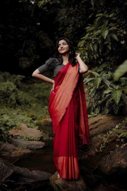 A woman in a vibrant red sari standing confidently in a lush forest setting.