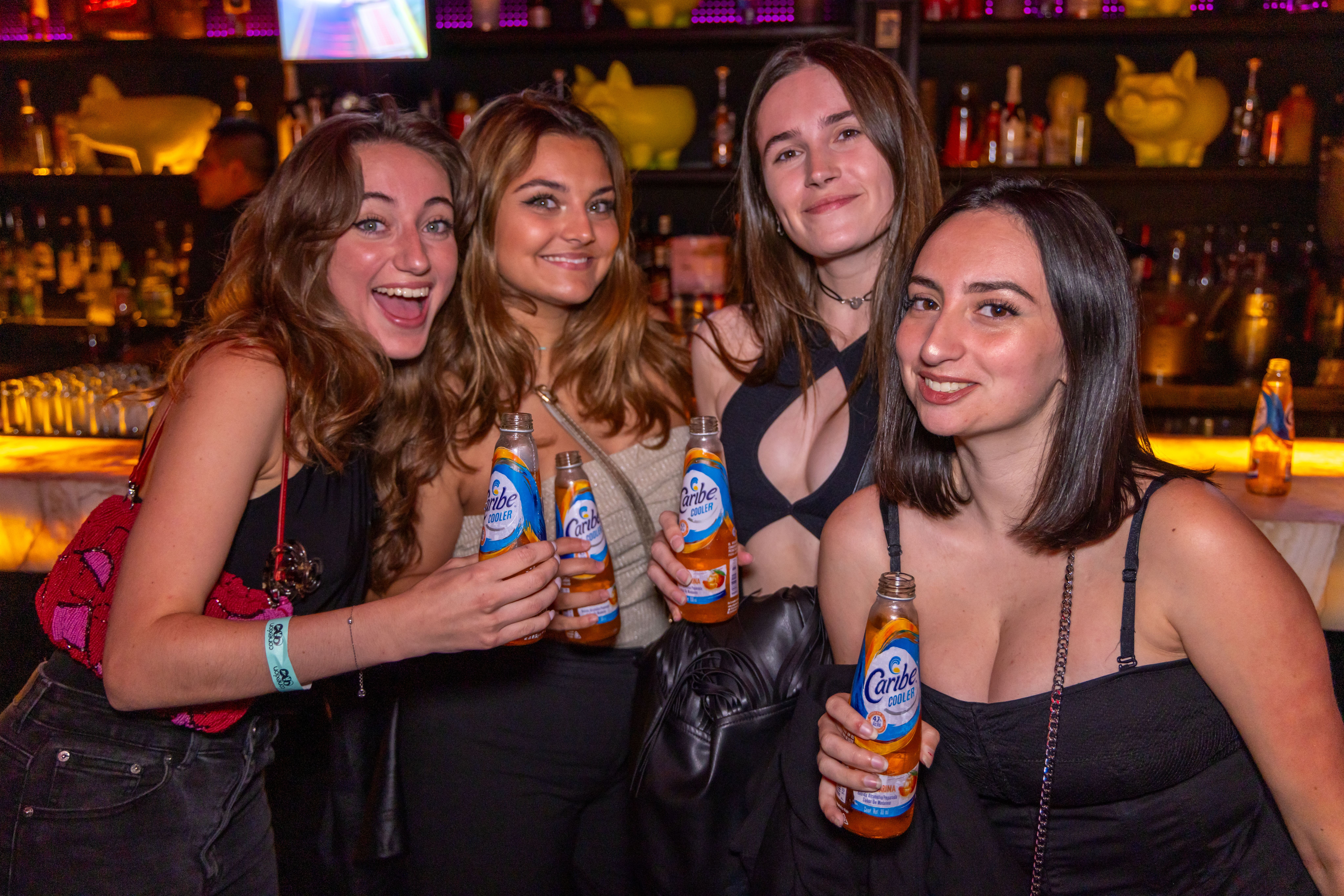 Four young women holding drinks and smiling in a lively bar atmosphere.