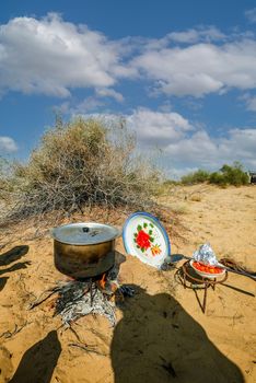 Rustic outdoor cooking setup in a desert landscape with a pot on a campfire, plate, and cooking tools.