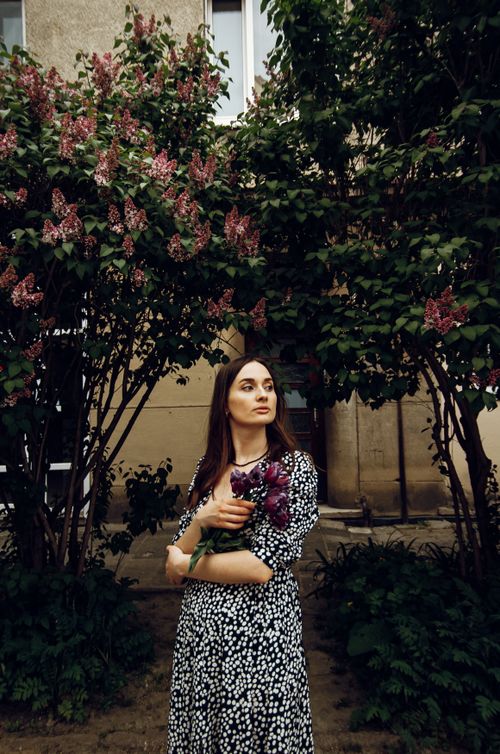 Free Caucasian woman holding flowers in front of a building and lush greenery. Stock Photo