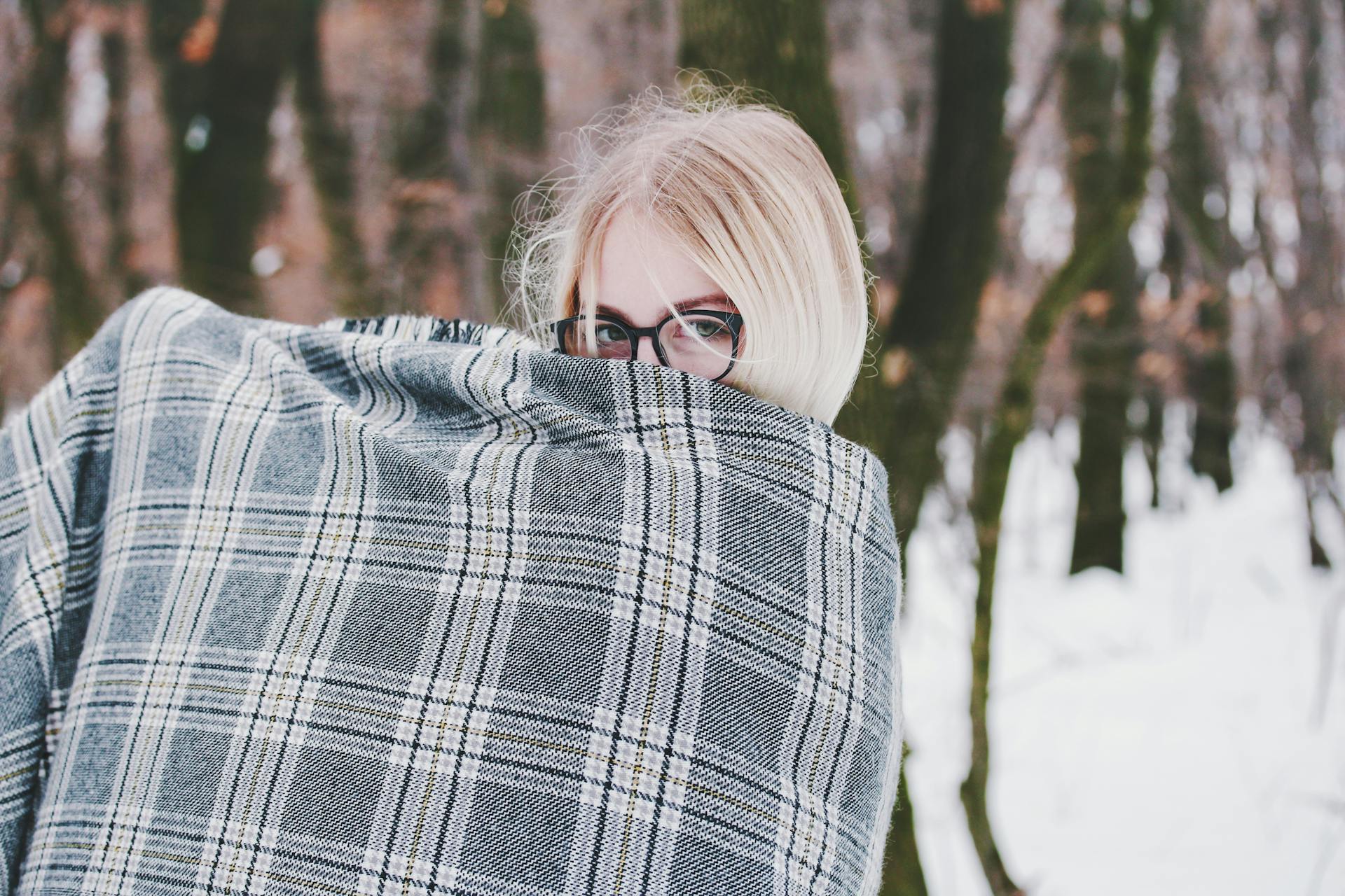 Woman with glasses wrapped in blanket in snowy forest during winter.