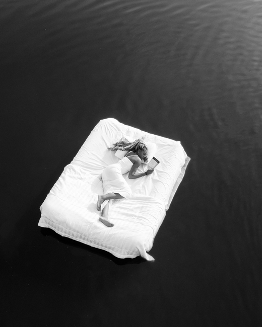 Black and white image of a woman reading on a floating mattress in water, top view.