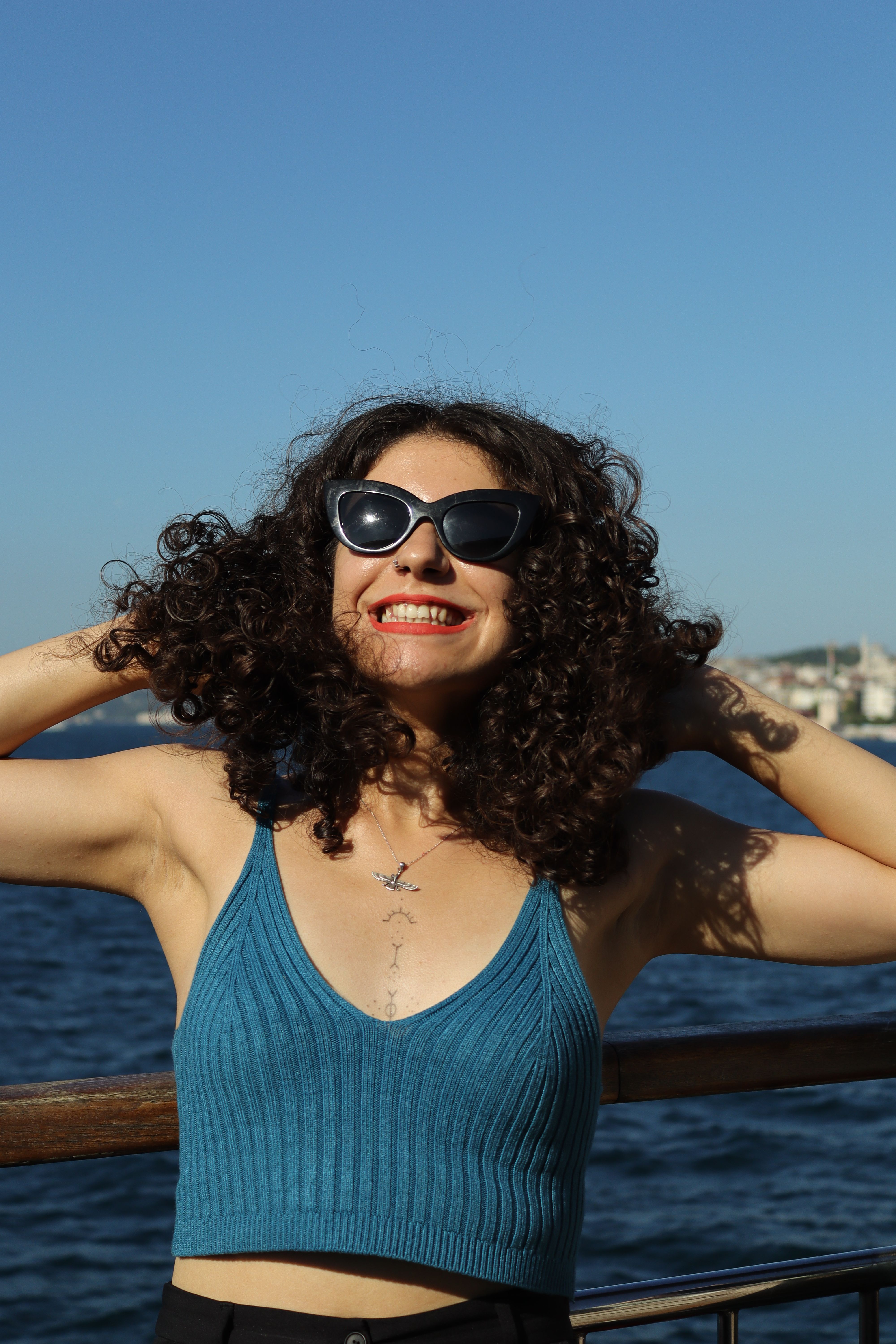 A smiling woman in sunglasses embraces the sunshine by the sea, embodying the essence of summer fun.