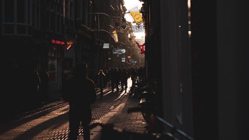 Free Silhouetted pedestrians walk a cobblestone street during sunset, embracing urban charm and vibrant lights. Stock Photo