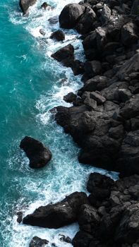 Aerial view of turquoise waves crashing on rugged black rocks along a scenic seashore.