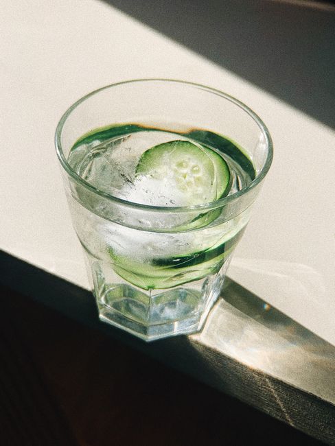 A clear glass of refreshing cucumber water with ice cubes glistening under sunlight on a table.