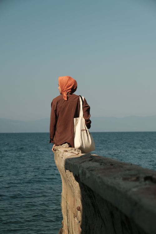 Free Woman in hijab sitting on a wall by the sea, gazing into the horizon. Peaceful and thoughtful moment. Stock Photo