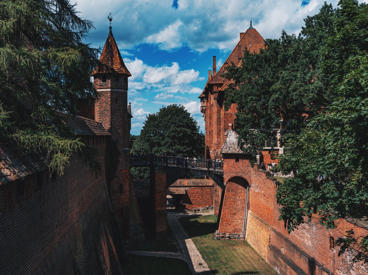 Visitors on a wooden bridge over a dry, grassy ditch, between a round brick tower with a pointed red roof and a brick arch.