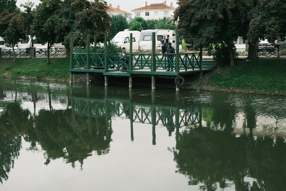 Peaceful park lake with a green pier, trees, and reflection. Three adults are visible.