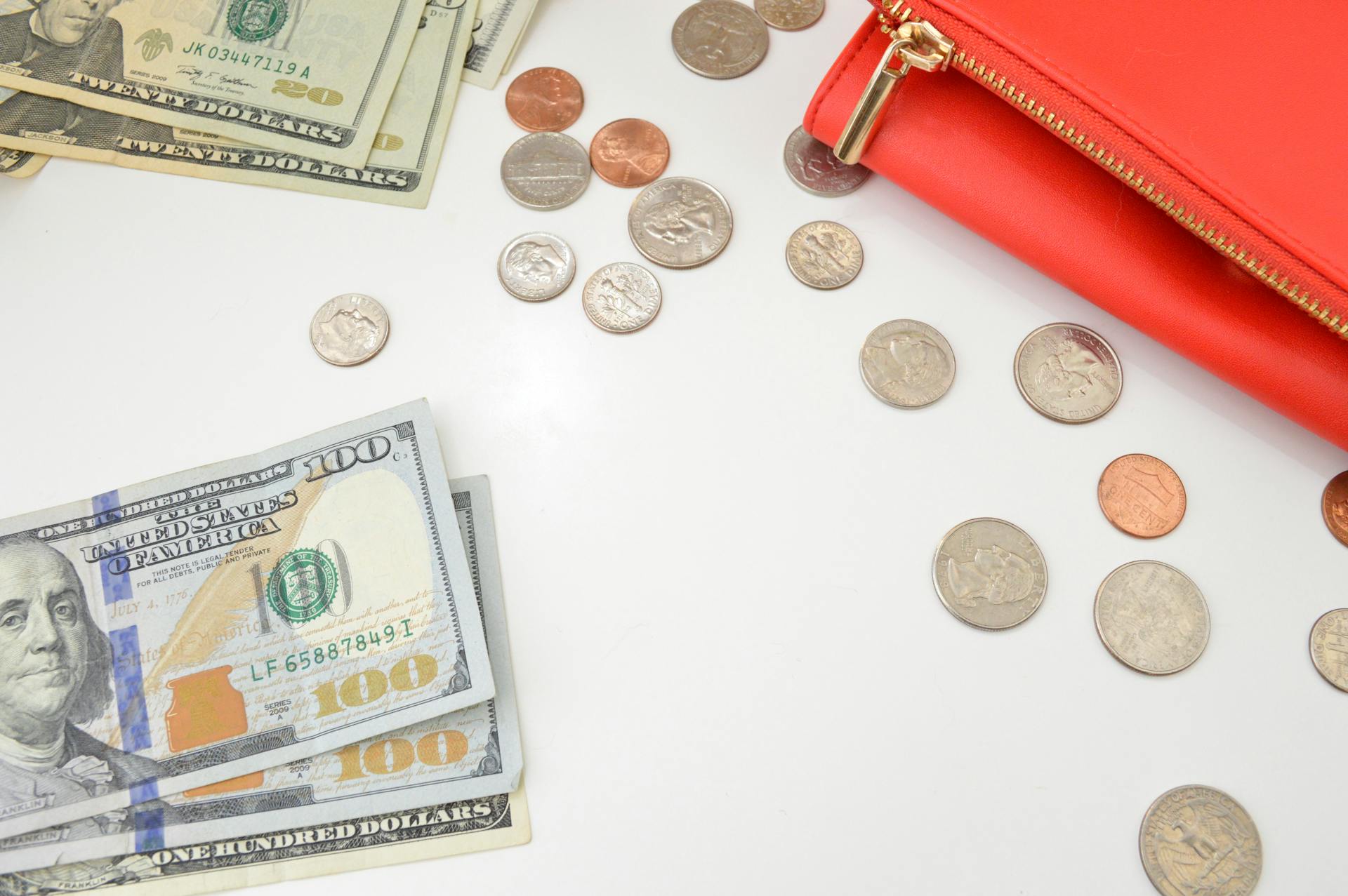 A top-view shot of scattered US currency notes and coins alongside a red wallet on a white surface.