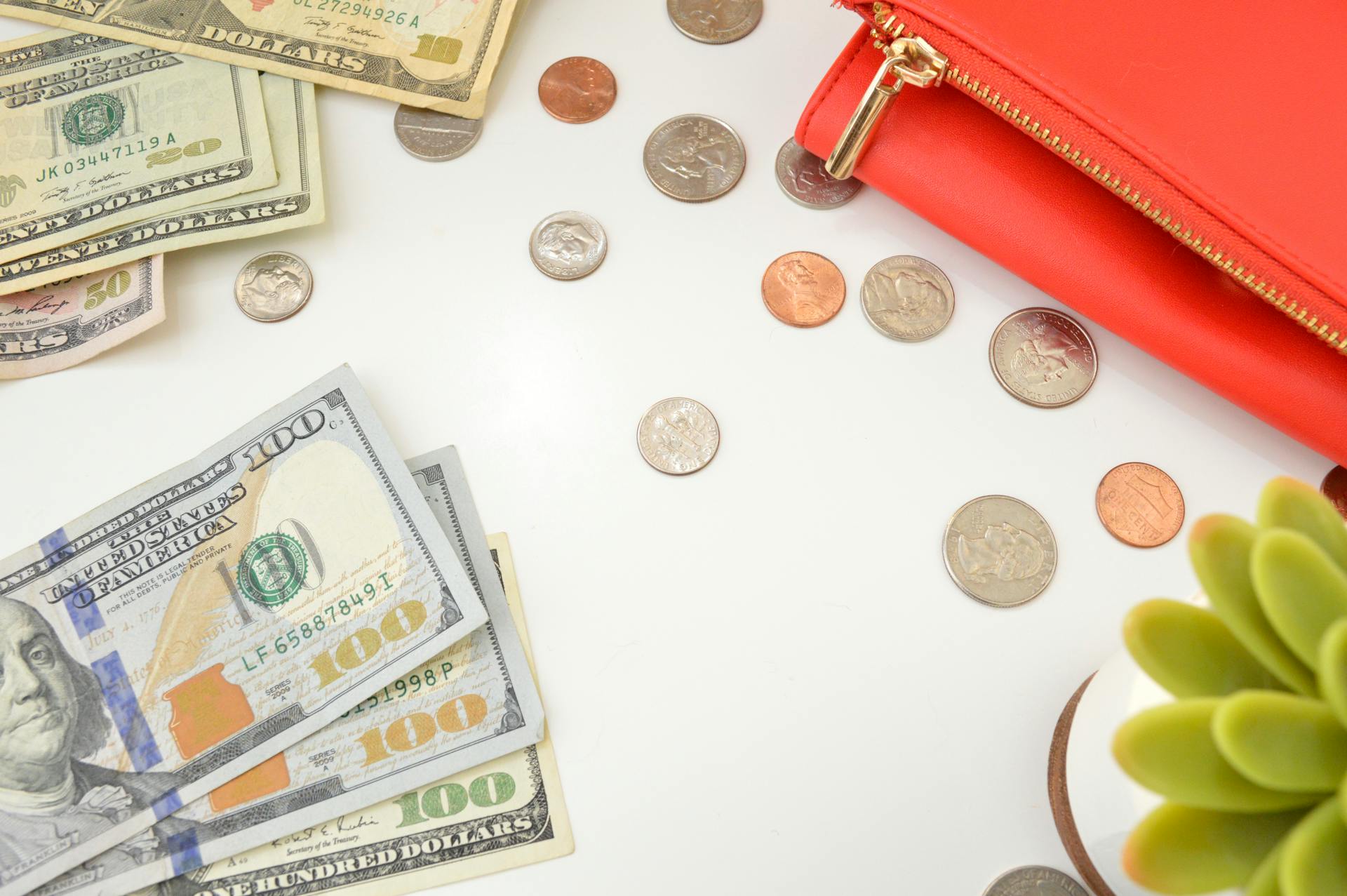 Dollar bills and change scattered near a red wallet on a white surface with a plant accent.