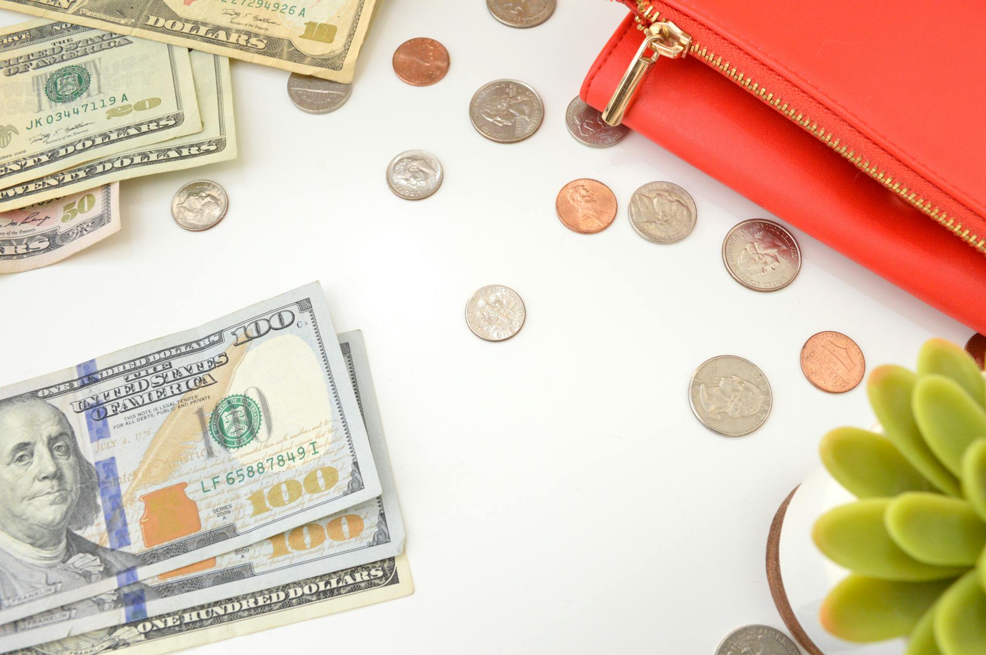 A top view of US dollar bills and coins scattered with a red wallet and succulent plant.