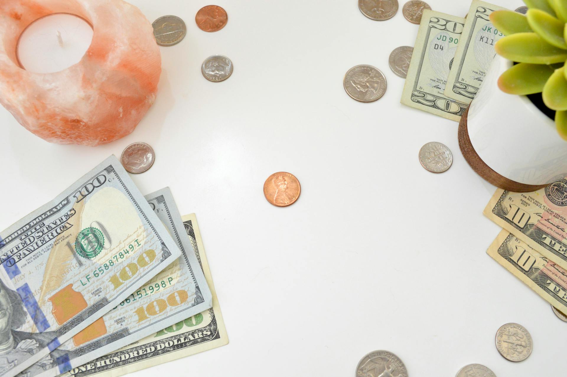 A minimalist flat lay of US currency, coins, a candle, and a plant on a white surface.