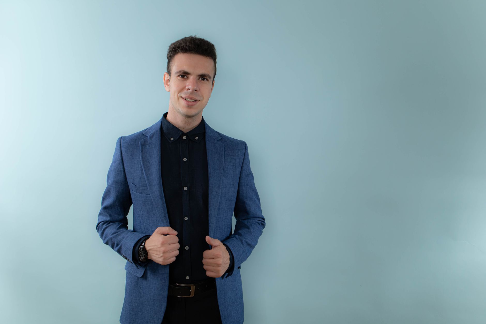 Stylish man posing against a light background, wearing a blue suit and a black shirt.