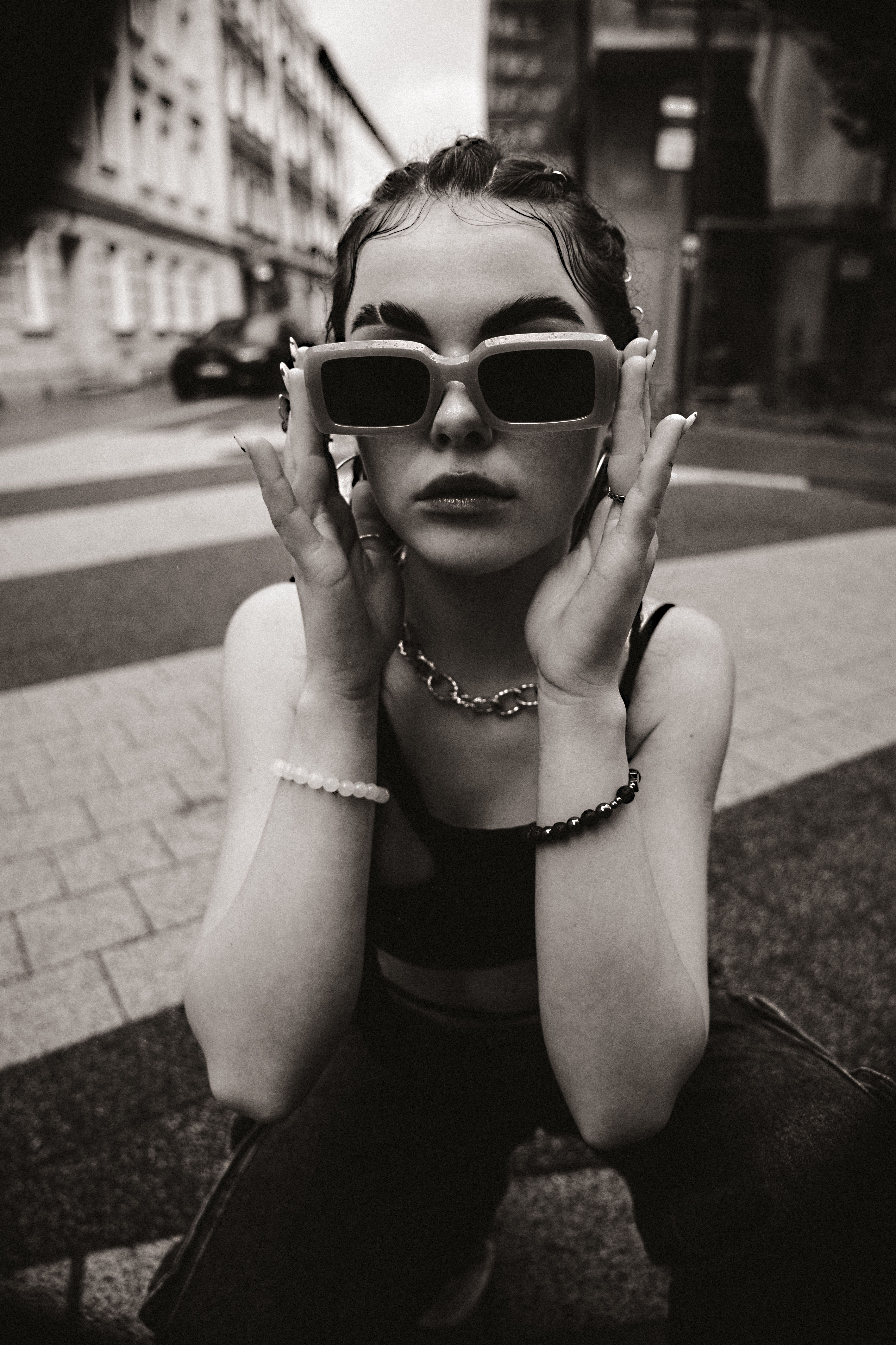 Stylish young woman crouching on city street wearing thick-rimmed sunglasses.