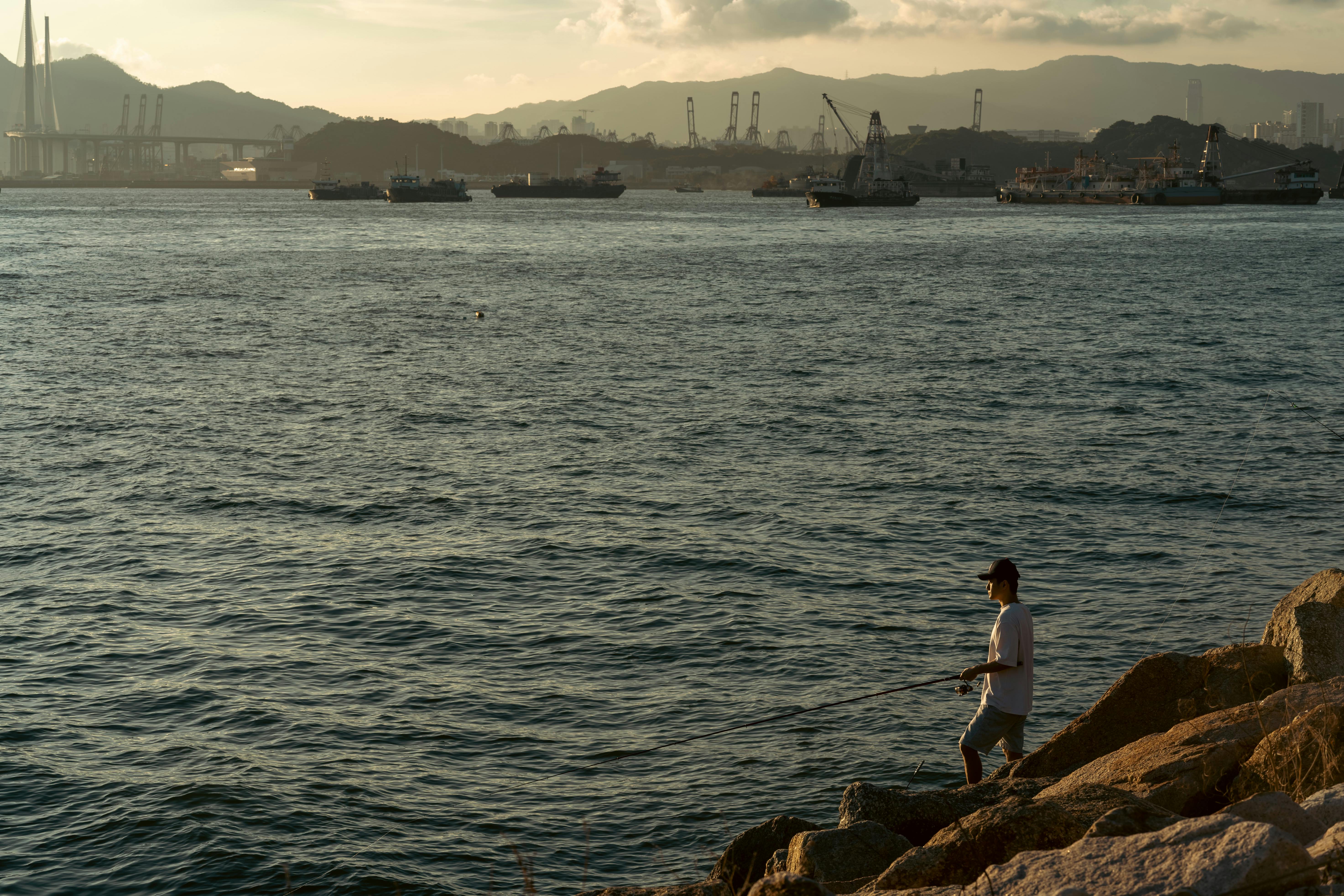 A lone man fishing by the rocky shore with an industrial harbor in the background during sunset.