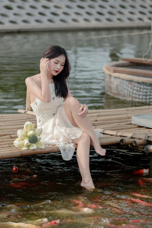 Young Woman in a White Dress Sitting with Her Foot in the Pond with Koi Fish 
