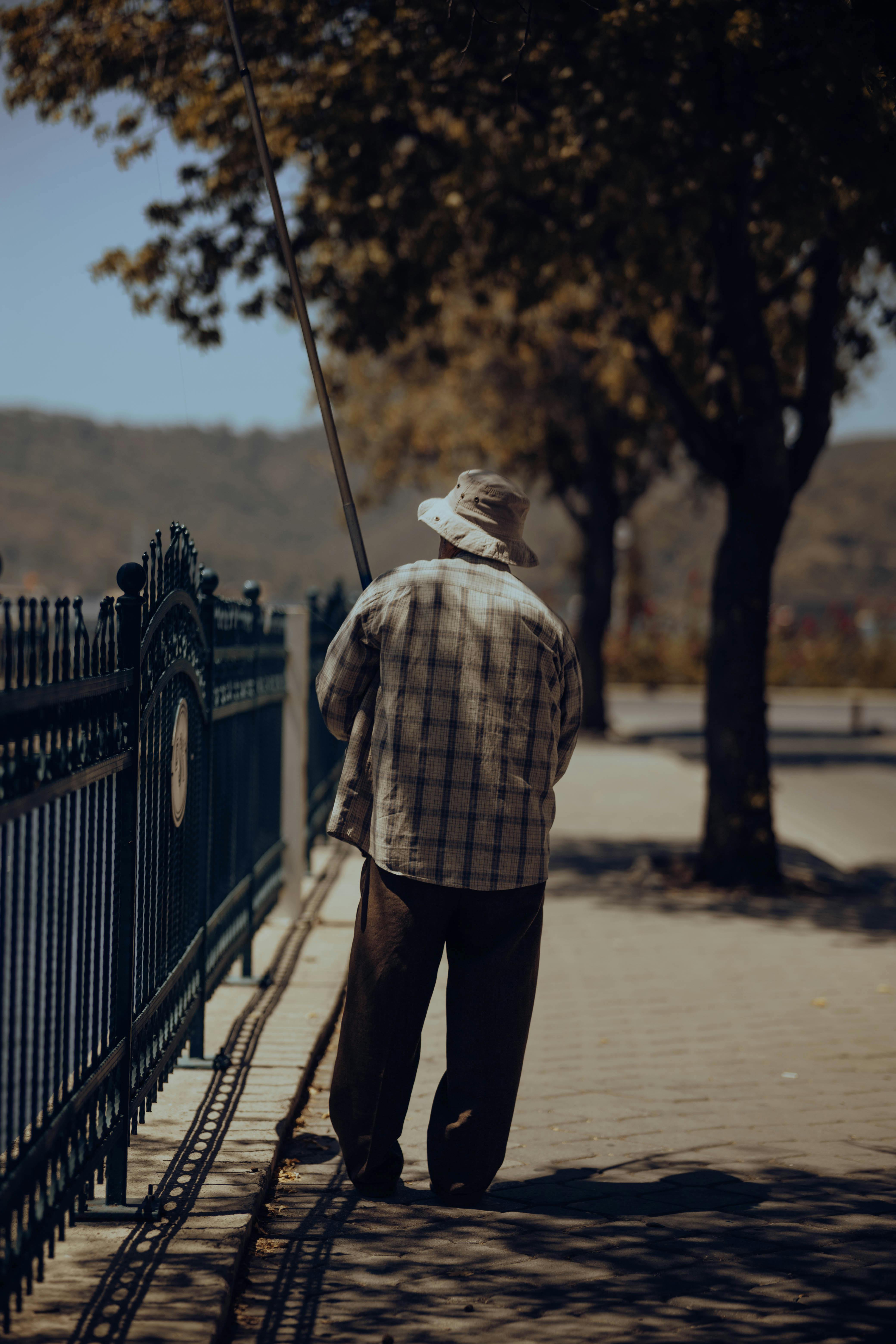 An elderly man in a hat fishing by a riverside fence on a sunny day.