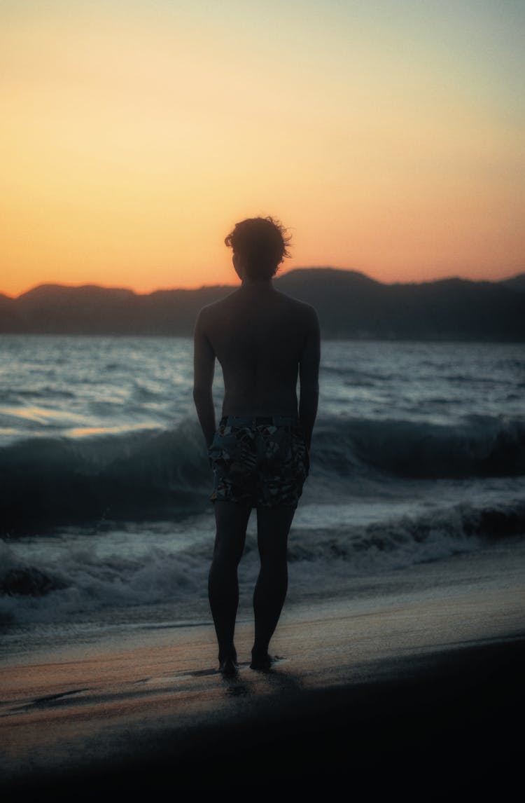 Young Man In Shorts Standing On A Beach At Sunset