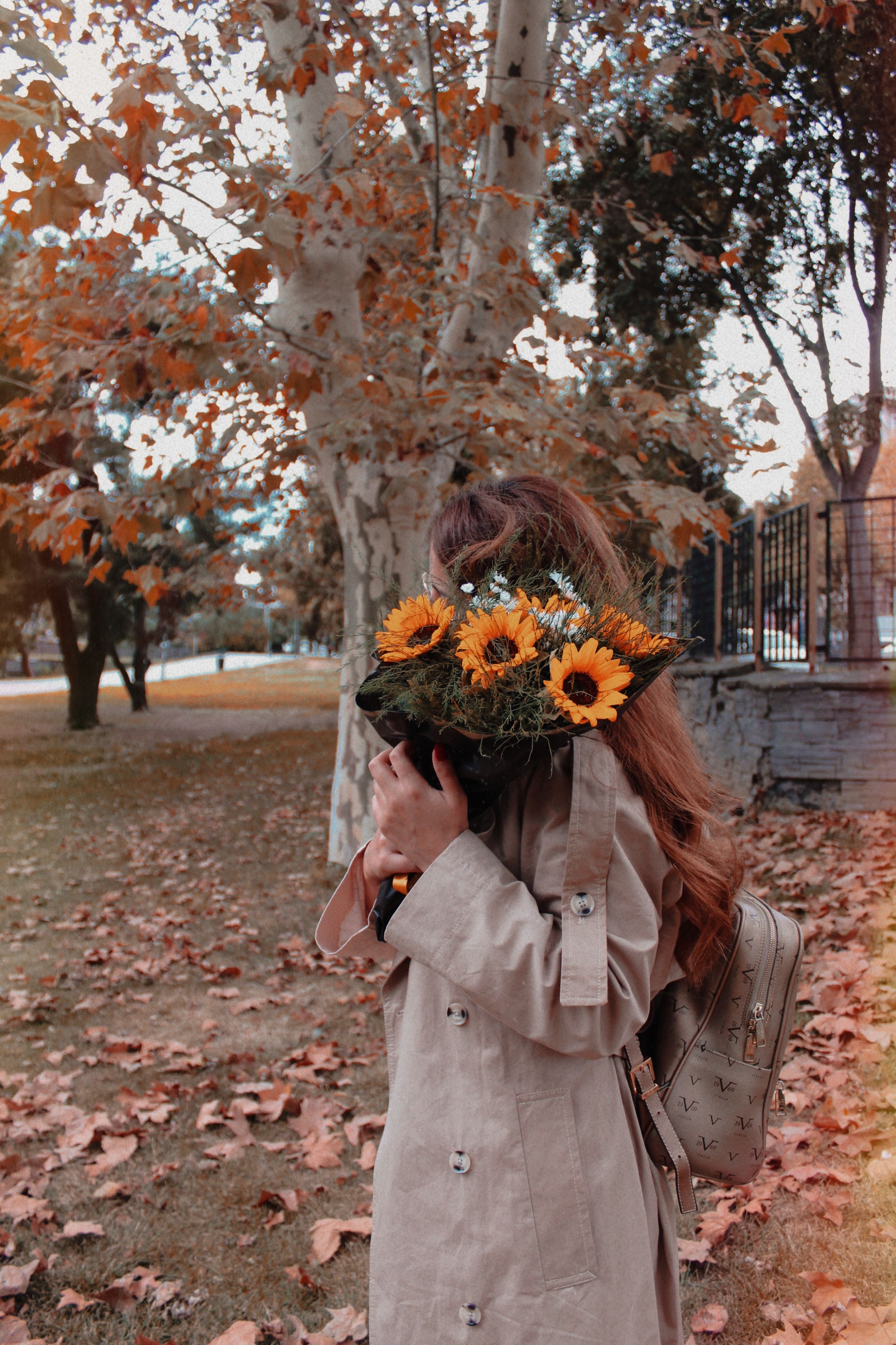 A woman in a beige coat holds sunflowers while standing in an autumn park.