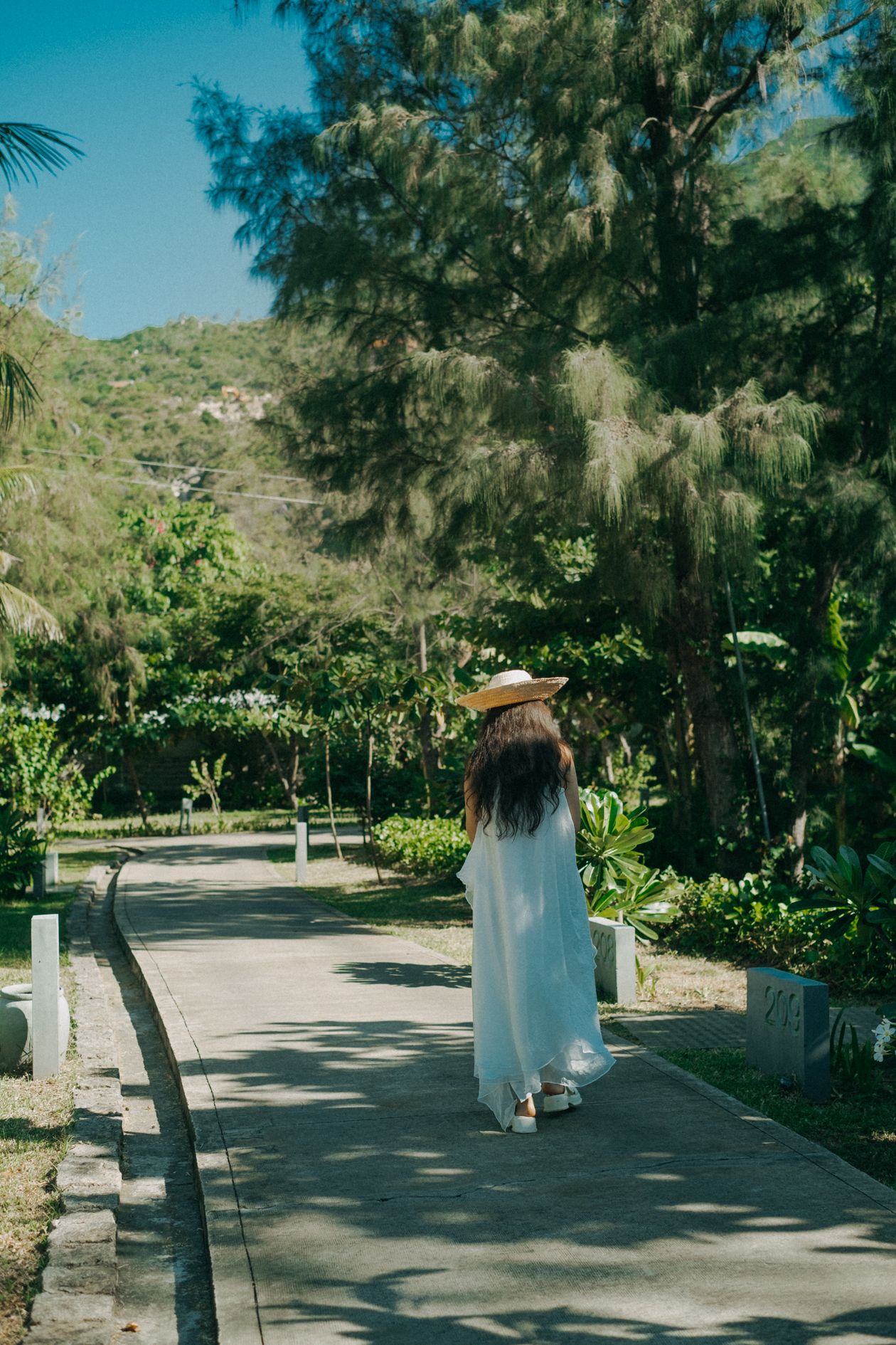 Woman from behind wearing a white flowy maxi dress and straw sunhat on a park path