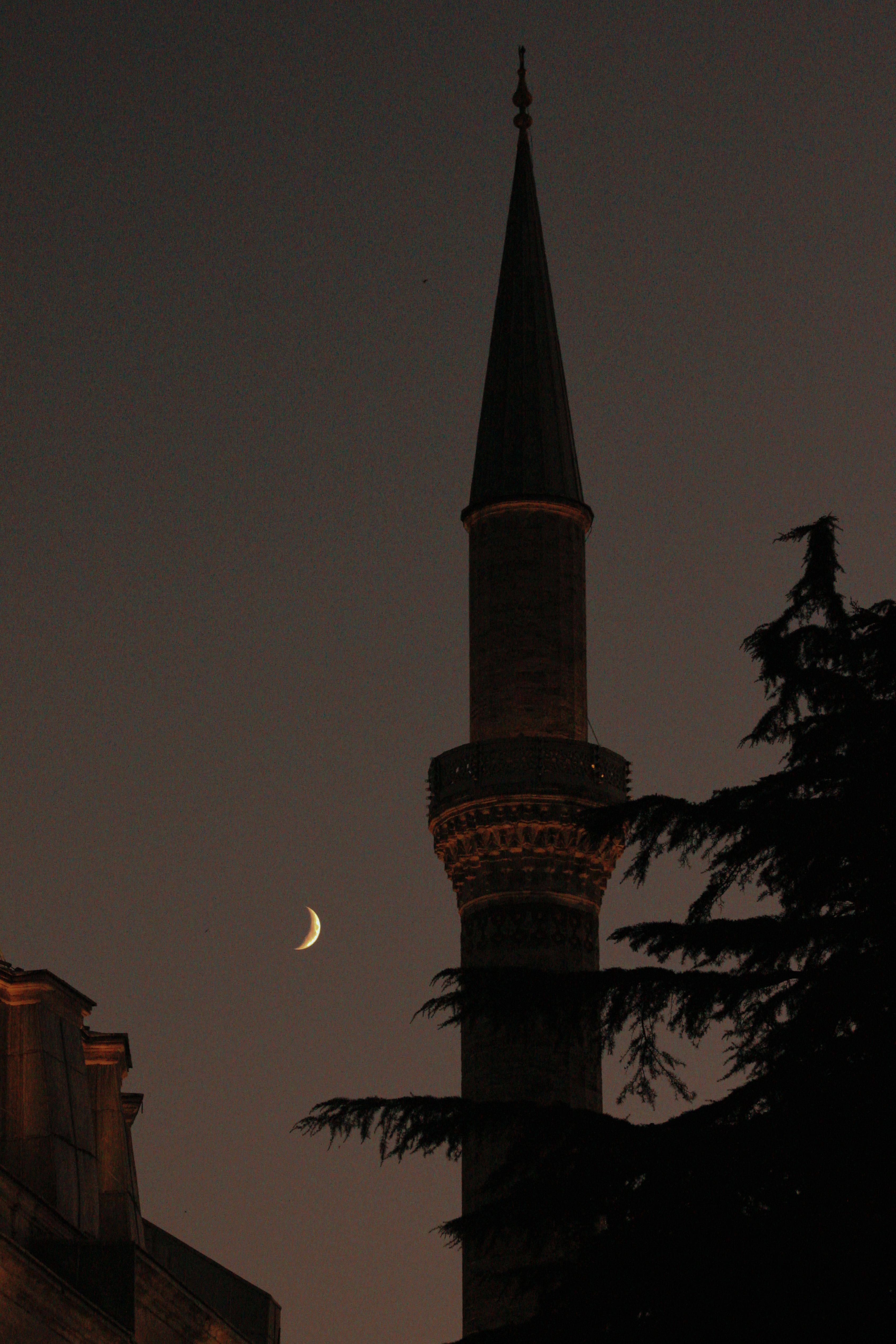 Silhouette of a mosque minaret under a crescent moon, captured at night, showcasing its architectural beauty.