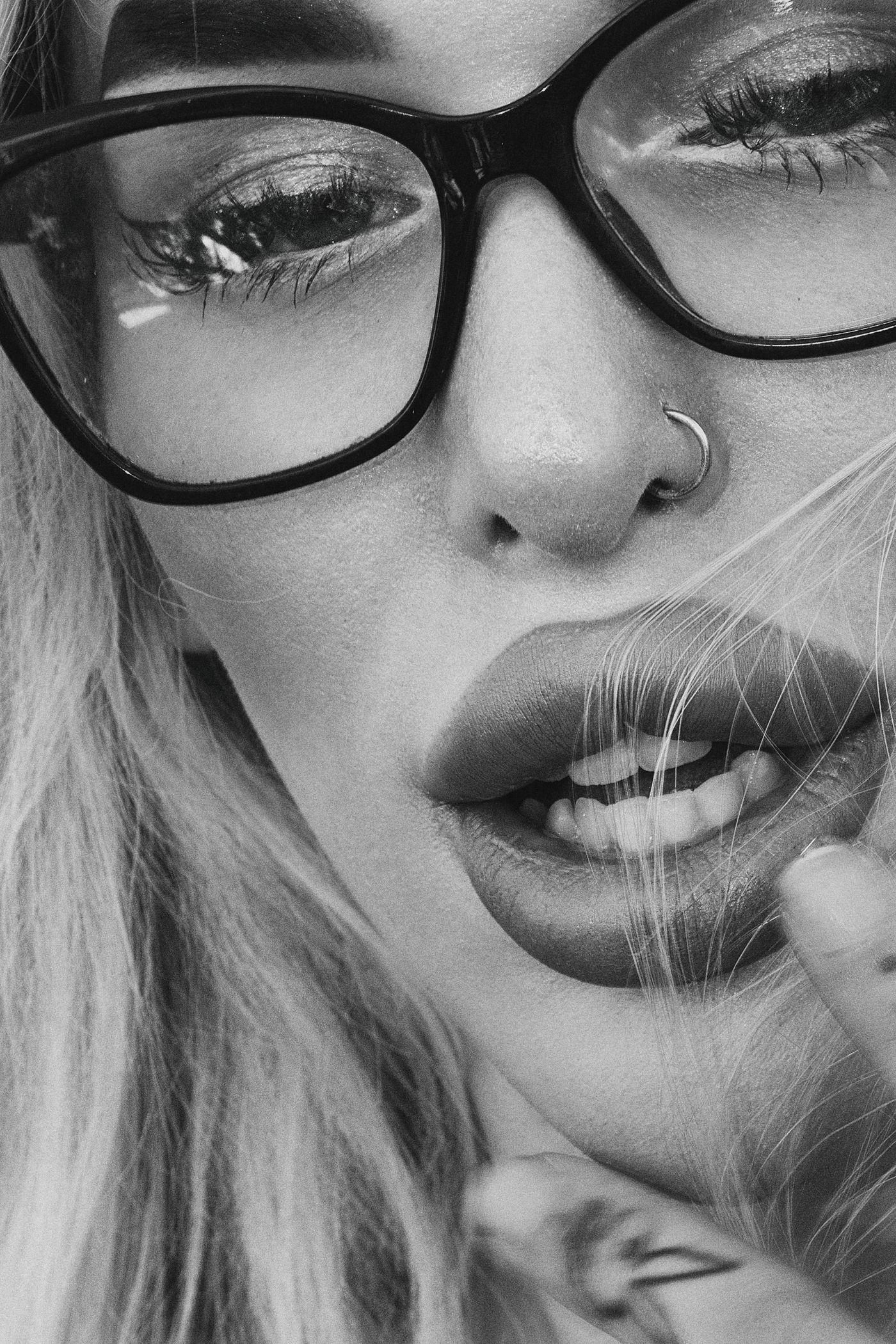 Black-and-white close-up of a woman wearing glasses and a nose ring, with closed eyes, visible eyelashes, and slightly parted natural-looking lips.