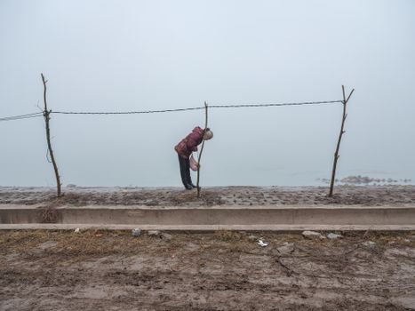 Elderly woman leans on makeshift fence by foggy lakeside in winter.