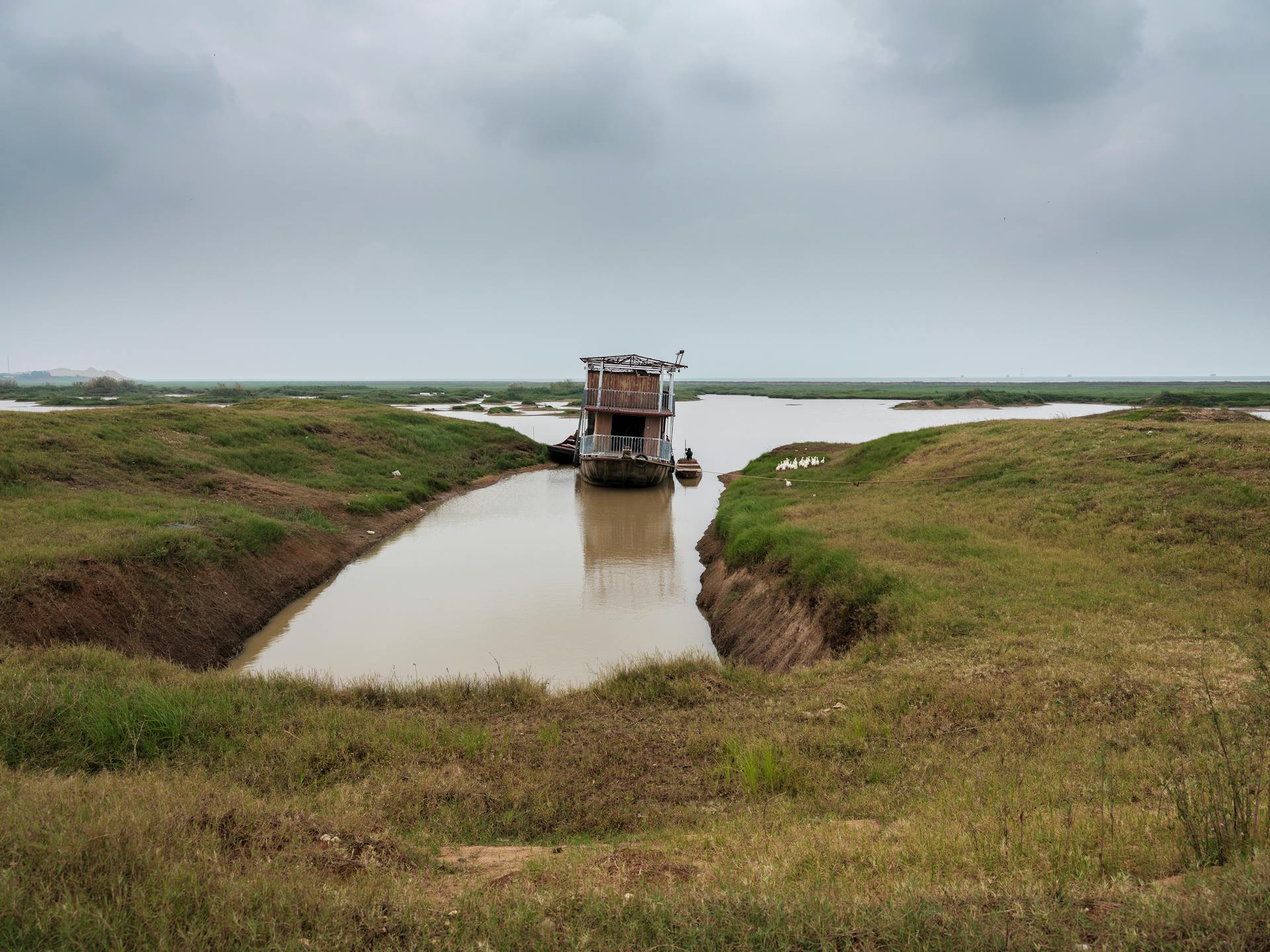 A tranquil scene of a boat anchored in a rural waterway, surrounded by grassy banks and an overcast sky.