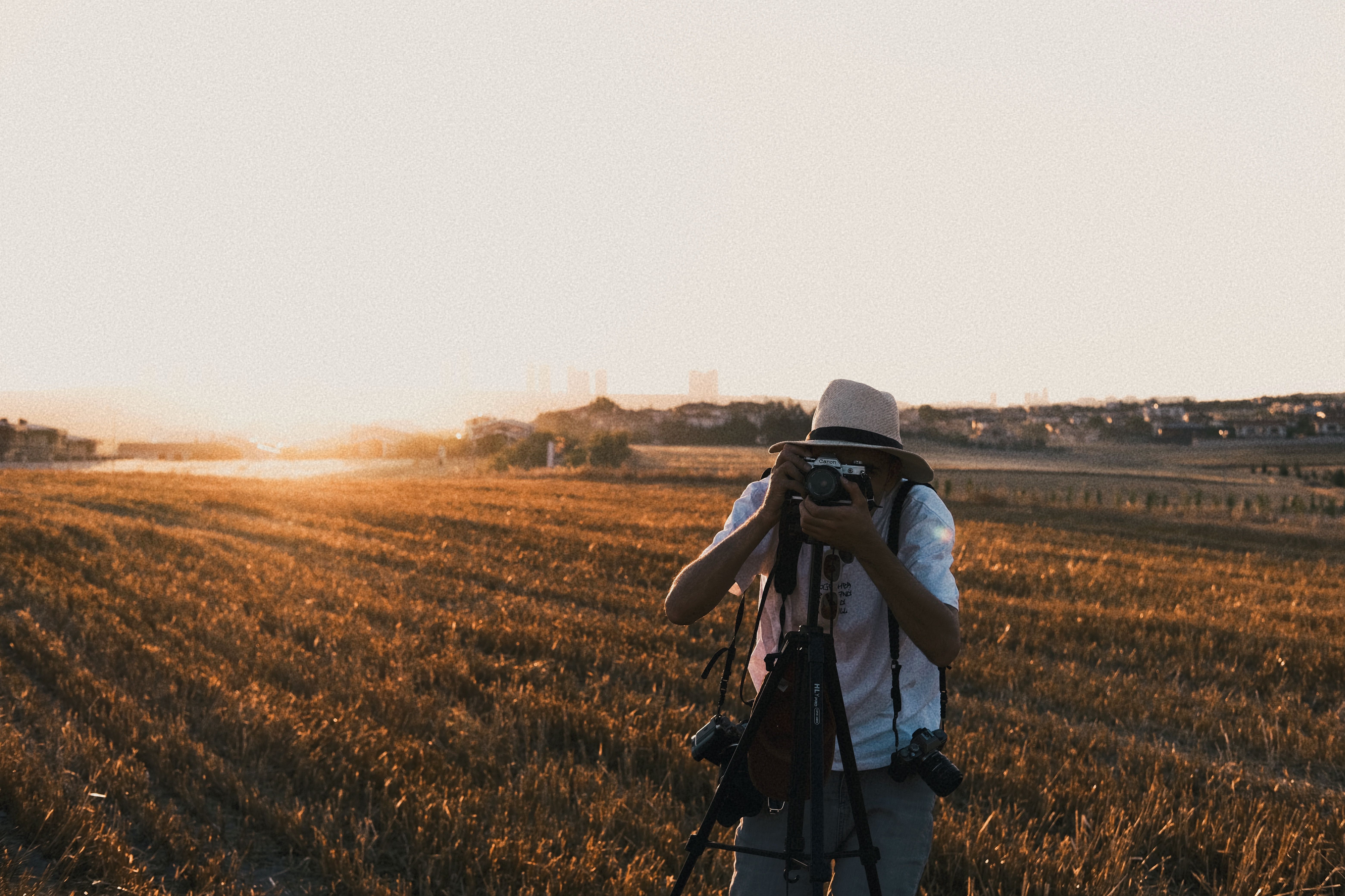 A photographer in a hat takes pictures in a sunlit rural field during sunset.